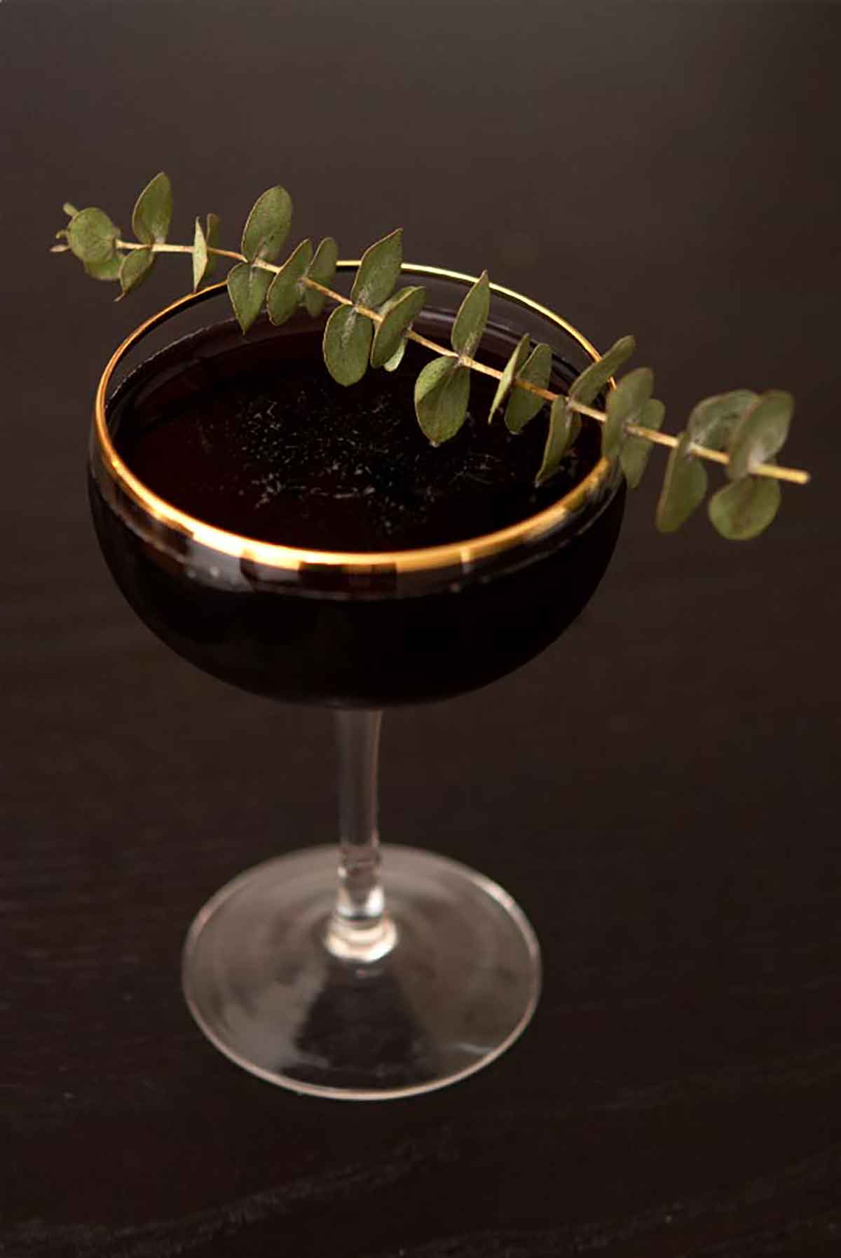 A black cocktail in a glass rimmed glass, garnished with eucalyptus.