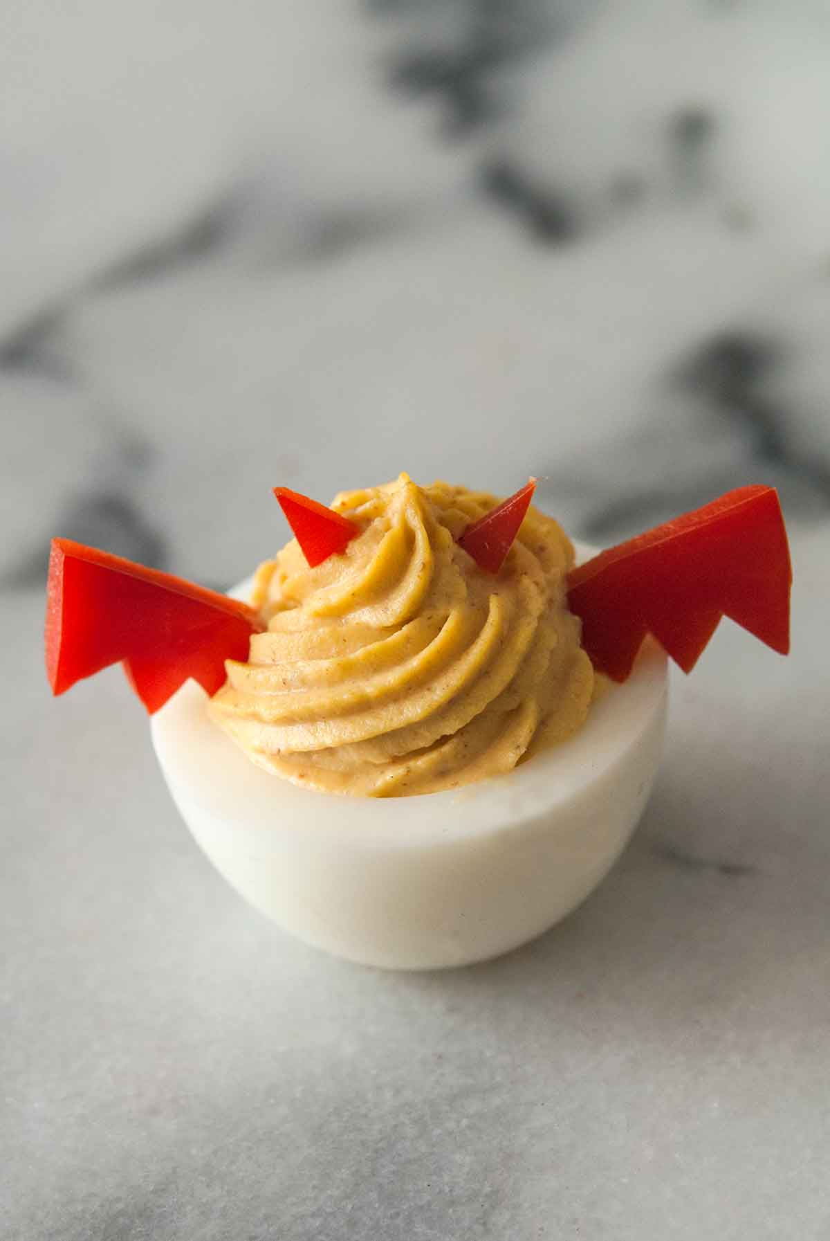 One deviled egg garnished with horns and wings made of red pepper on a marble board.