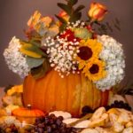 A pumpkin full of flowers on a table, surrounded by bread, cheese, fruit and vegetables.