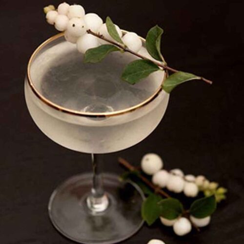 A cocktail garnished with ghost berries on a black table with berries at its base, and in the background.