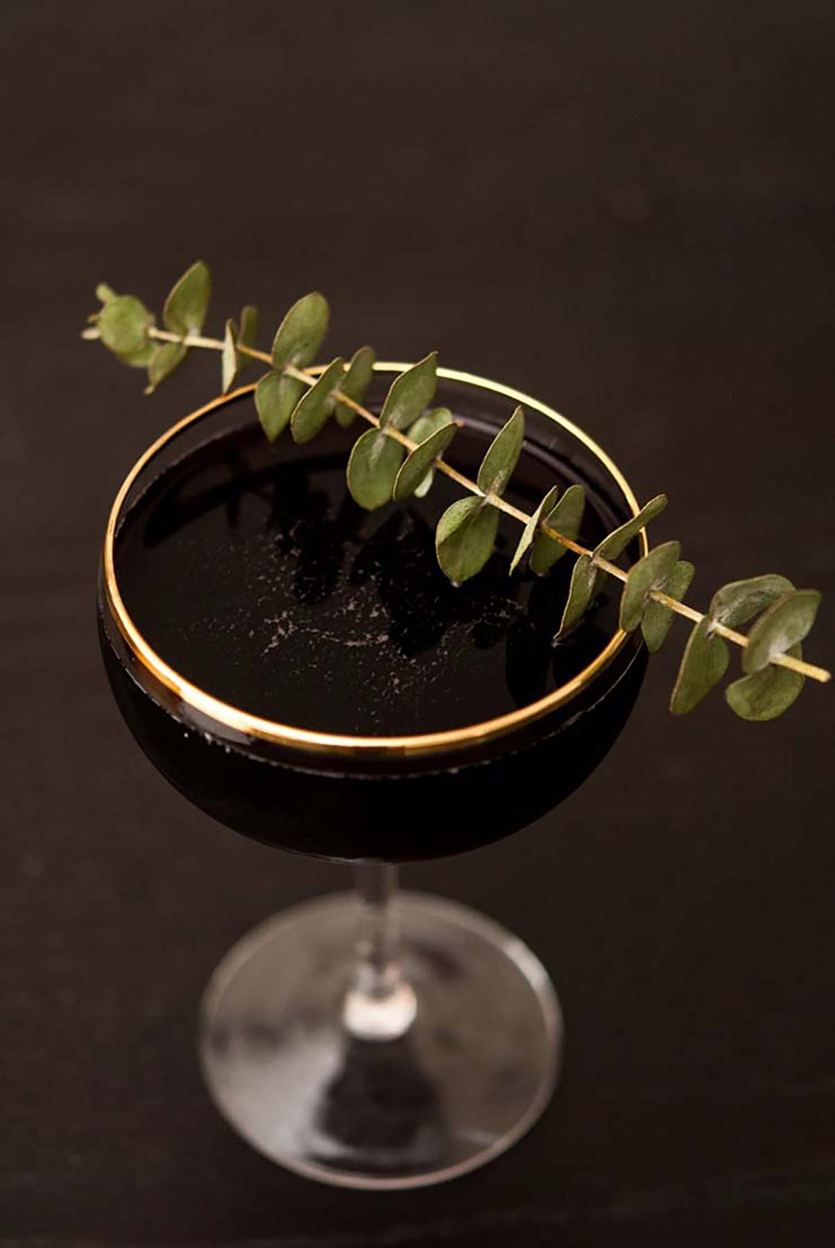 A cocktail in a gold-rimmed glass on a dark table, garnished with a single stem of eucalyptus. 