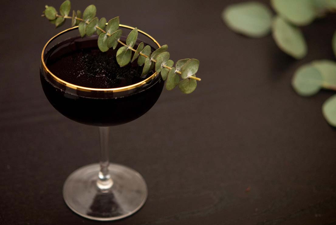 A cocktail in a gold-rimmed glass, garnished with eucalyptus on a dark table.