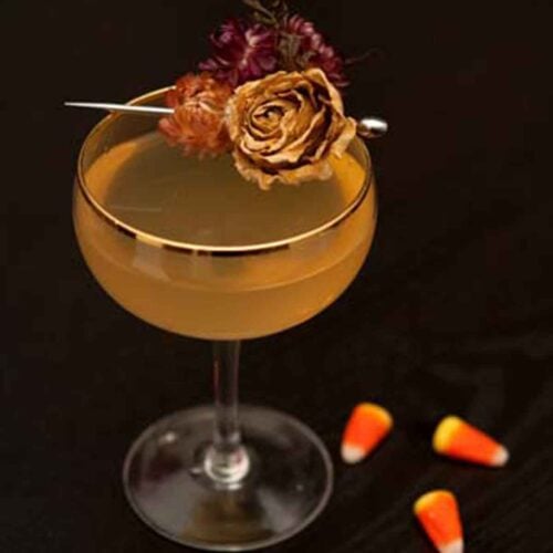 A cocktail on a table garnished with dry flowers with 3 candy corns at its base.