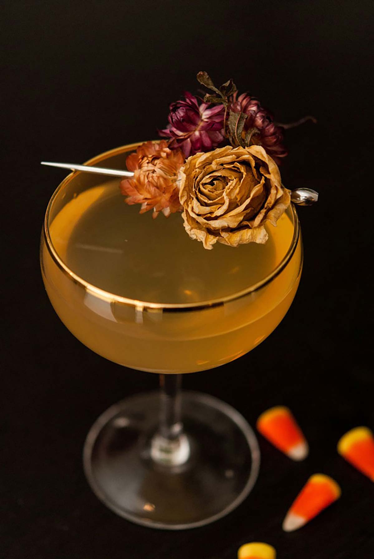 A cocktail garnished with dry flowers on a black table sprinkled with 3 candy corns.