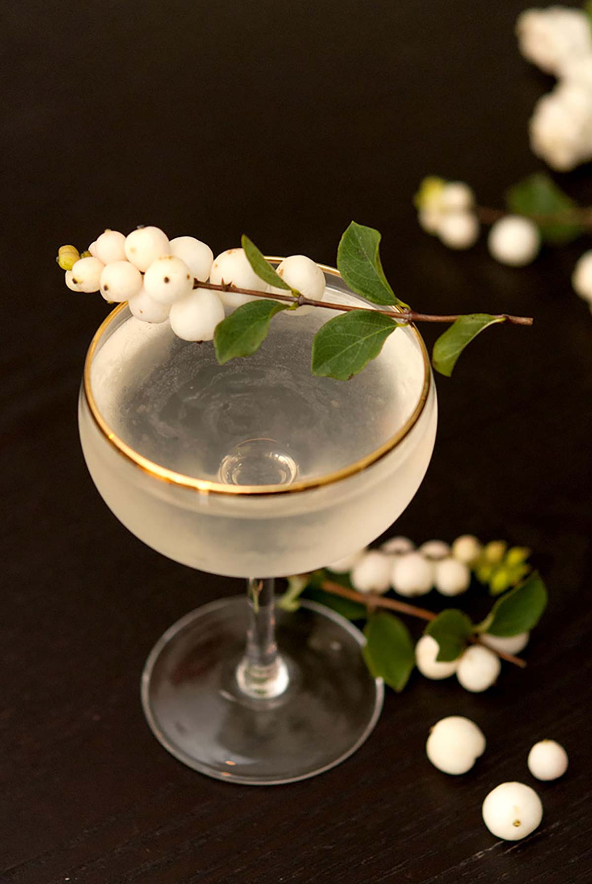 A clear cocktail in a gold-rimmed glass, garnished with white ghost berries on a black table, sprinkled with a few loose ghost berries.