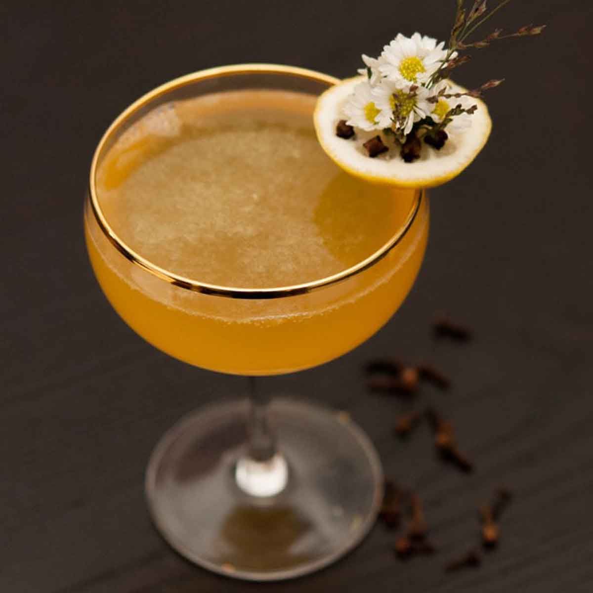 A cocktail garnished with lemon, flowers and cloves on a table, with lemon slices, flowers and cloves at its base.