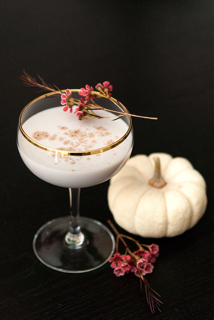 A cocktail in a coup glass, garnished with cinnamon and a sprig of flowers, with flowers at its base on a dark table.