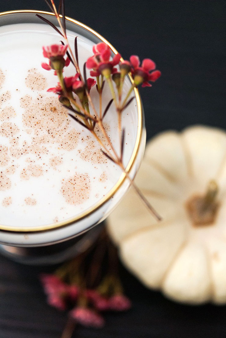 The top of a cocktail with a gold rim, garnished with spice and small flowers with a white pumpkin in the background.