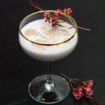 A cocktail in a coup glass, garnished with cinnamon and a sprig of flowers, with flowers at its base on a dark table.