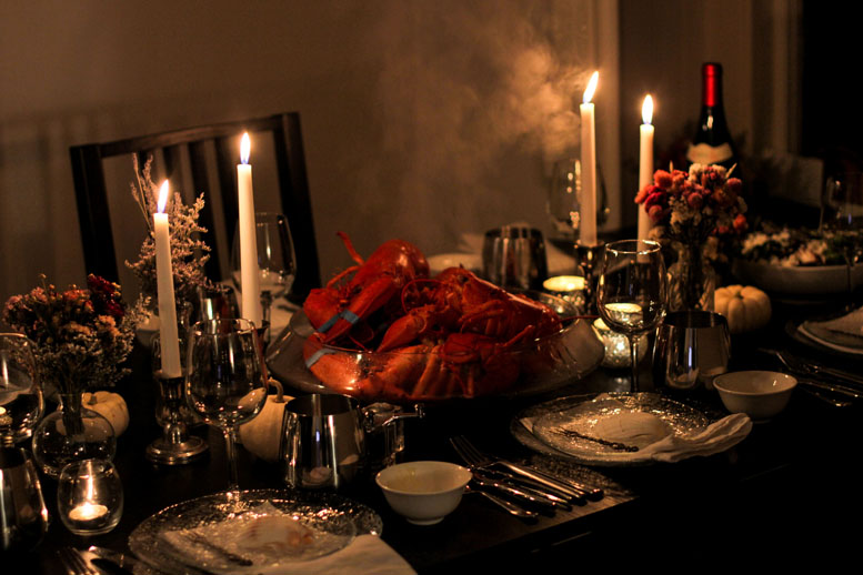 A large bowl of lobsters in the center of a table, surrounded by plates, small white bowls, cutlery, candles and red wine.