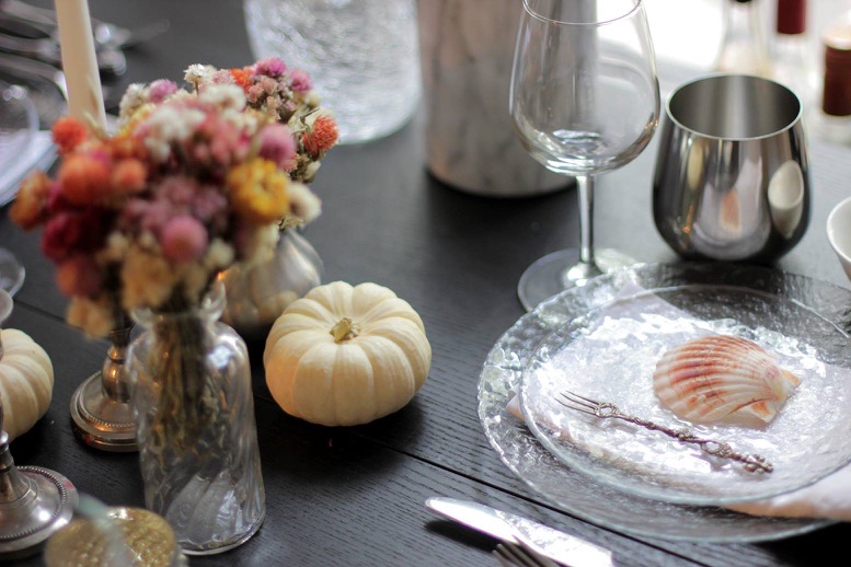 A table with white pumpkins, small vases with dry flowers, plates, silver cups and a shell placed in the center of a plate.