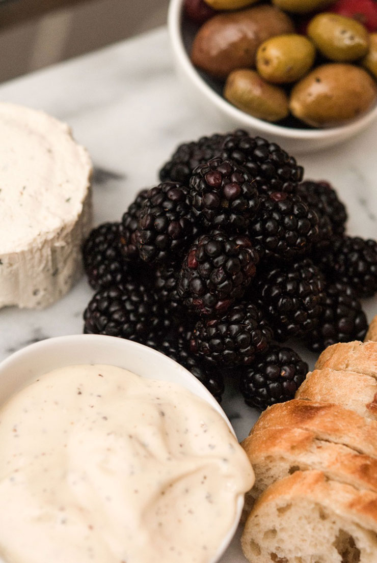 A pile of blackberries on a marble plate surrounded by sliced bread, cheese and a bowl of olives.