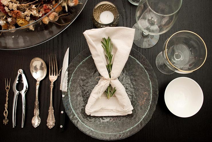 A napkin tied with string, with a sprig of rosemary tucked inside. The napkin is on top of a bowl, on top of a plate surrounded by utensils.