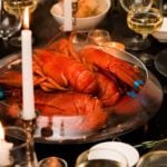 A large bowl with 5 cooked lobsters in the center of a table, set with glasses, candles and bowls of bread.