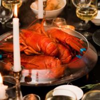 A large bowl with 5 cooked lobsters in the center of a table, set with glasses, candles and bowls of bread.