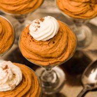 A glass of pumpkin cheesecake mouse, topped with whipped cream on a metal tray beside 4 others and a spoon.