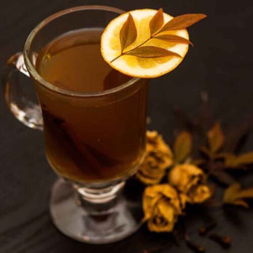A mug of hot toddy, garnished with an orange slice, topped with a small lead, with a few dry roses and leaves at its base.