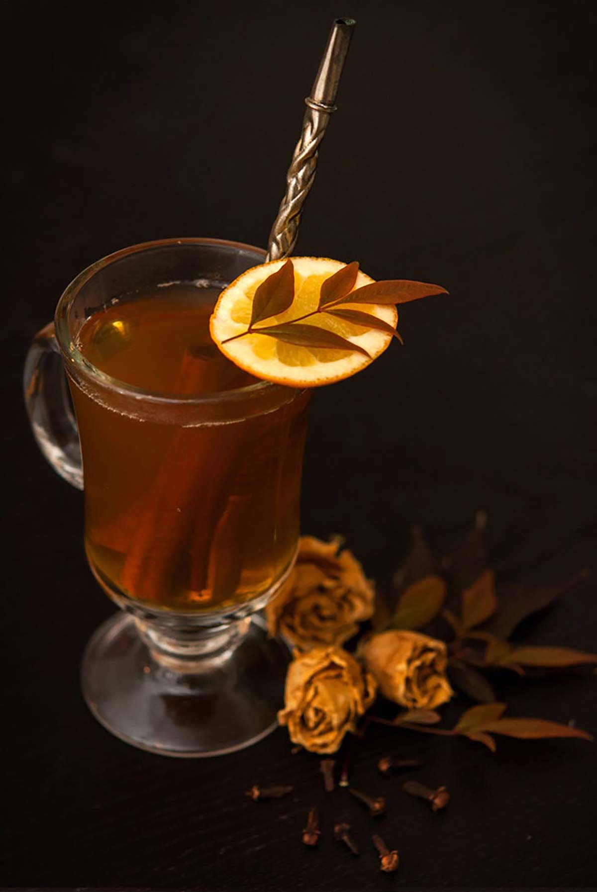 A Hot Toddy, with a tea infuser, garnished with an orange slice, with flowers at its base.