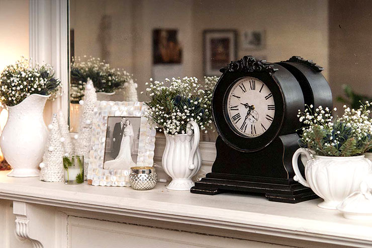 A mantle with a vintage wedding photo, a vintage clock and various vases holding baby's breath and greenery.