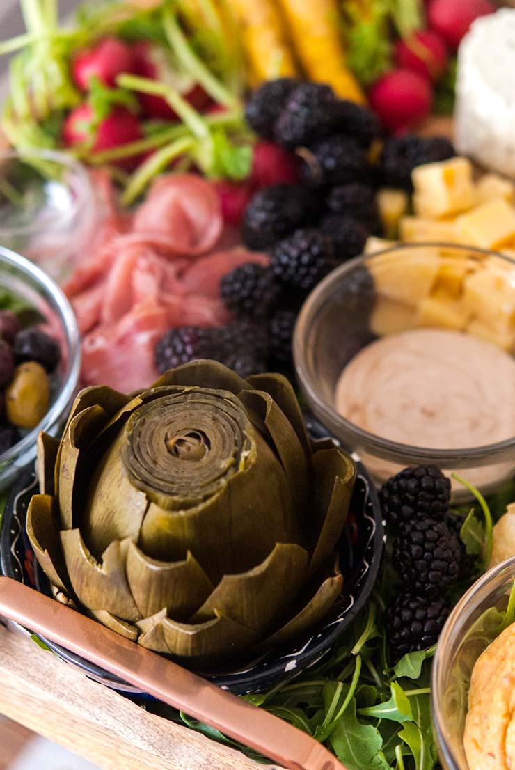 A tray with an artichoke, a bowl with dipping sauce, a bowl with olives, prosciutto, cheese blackberries and radishes.