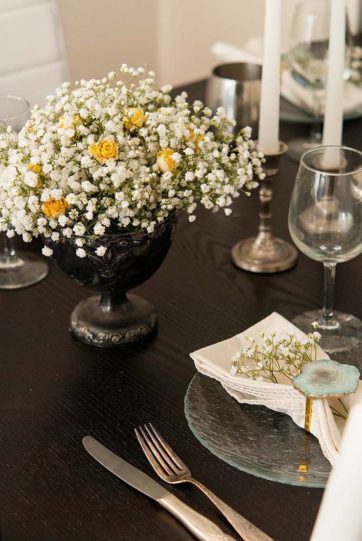 A dining room table set with plates, napkins, glasses, candles and a baby's breath bouquet with yellow roses.