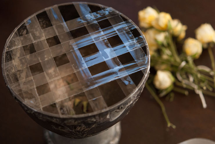 A tarnished silver bowl with tape criss-crossed on top to make a grid with yellow roses in the background on a table.