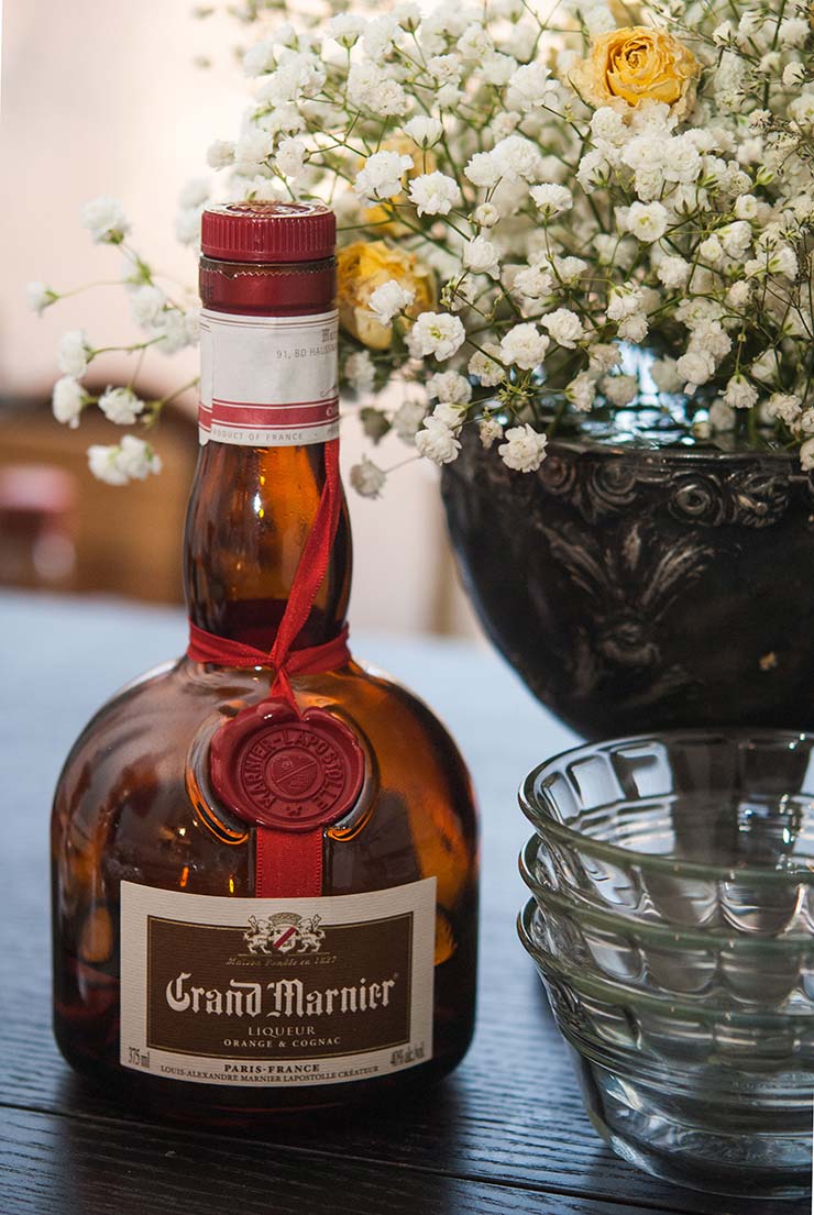 A bottle of Grand Marnier on a table next to 3 ramekins and bouquet of baby's breath and roses.