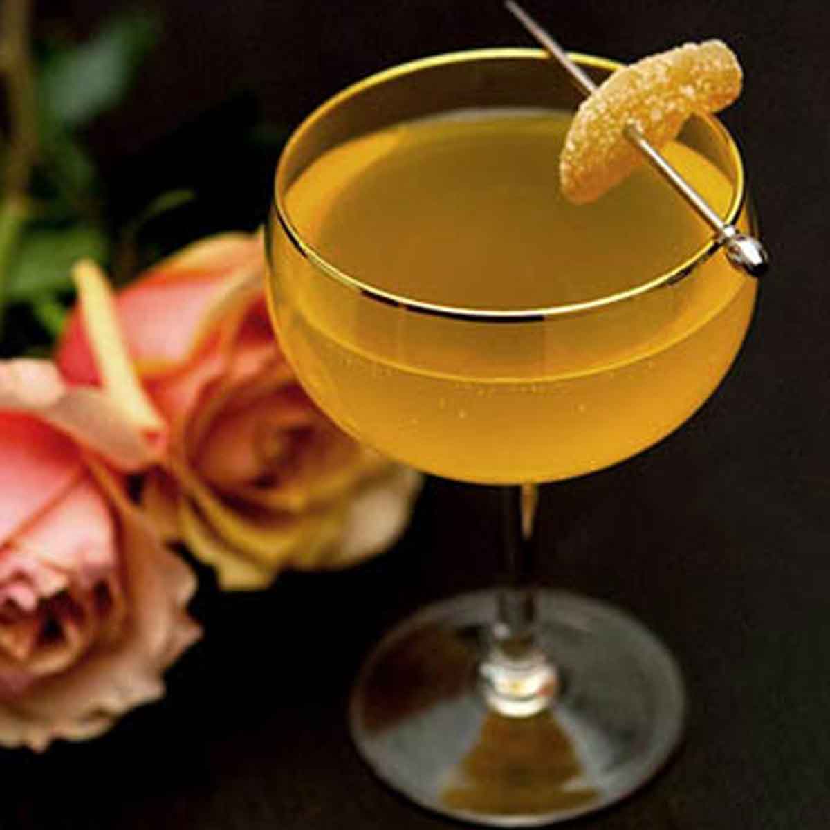 A cocktail garnished with a small slice of ginger on a dark table beside 2 roses.