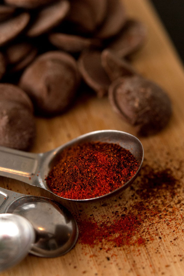 A measuring spoon with light and dark spices on a wooden cutting board beside a pile of chocolate.