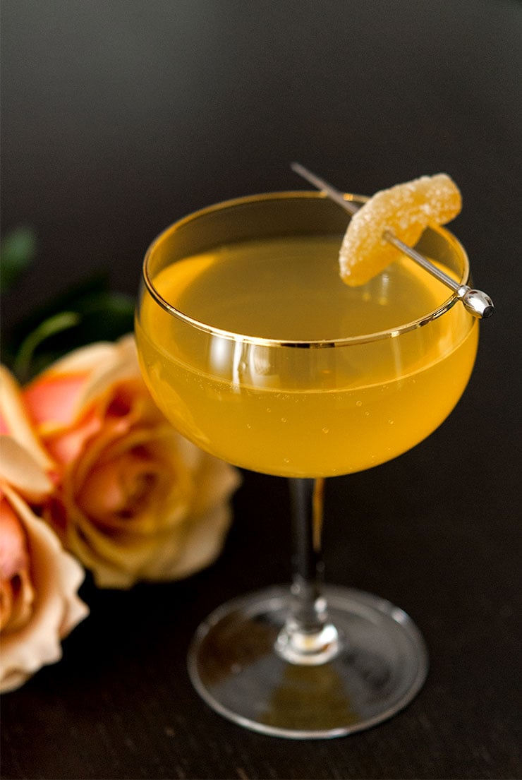 A cocktail on a black table garnished with ginger beside 2 roses.