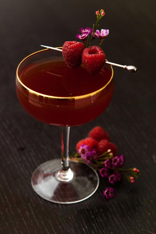 A cocktail on a dark table garnished with raspberries and small flowers, with a few raspberries and flowers at its base.