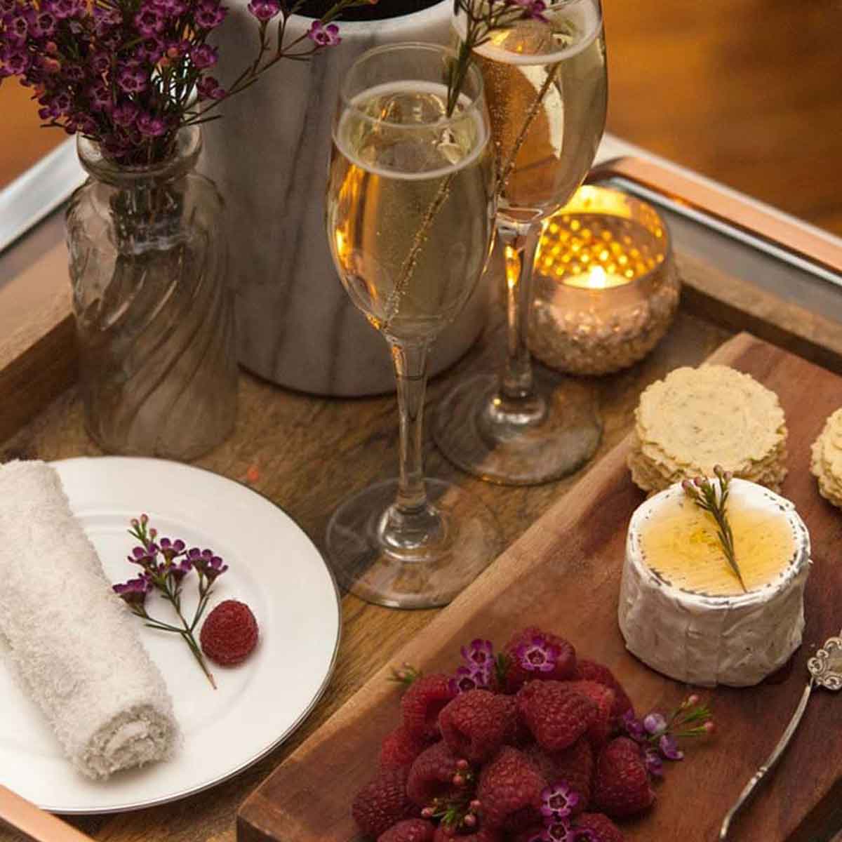 A wooden tray holding 2 champagne glasses with an assortment of cheeses, berries, crackers and flowers.