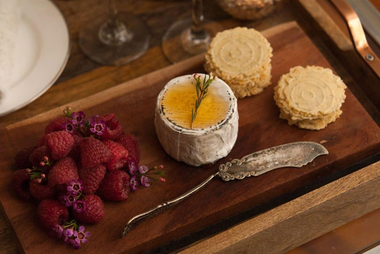 A cheeseboard with raspberries, cheese, garnished with honey and a sprig of flowers, an antique cheese knife and crackers.