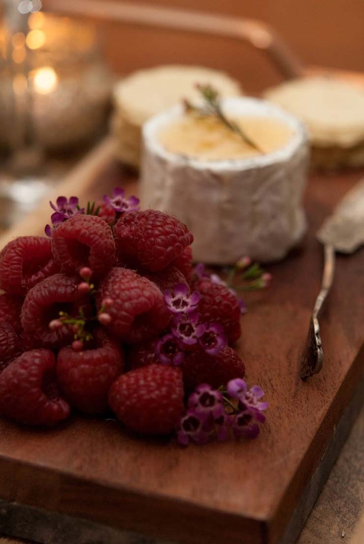 Raspberries stacked on a wooden cheeseboard, garnished with little flowers, beside cheese and a cheese knife.