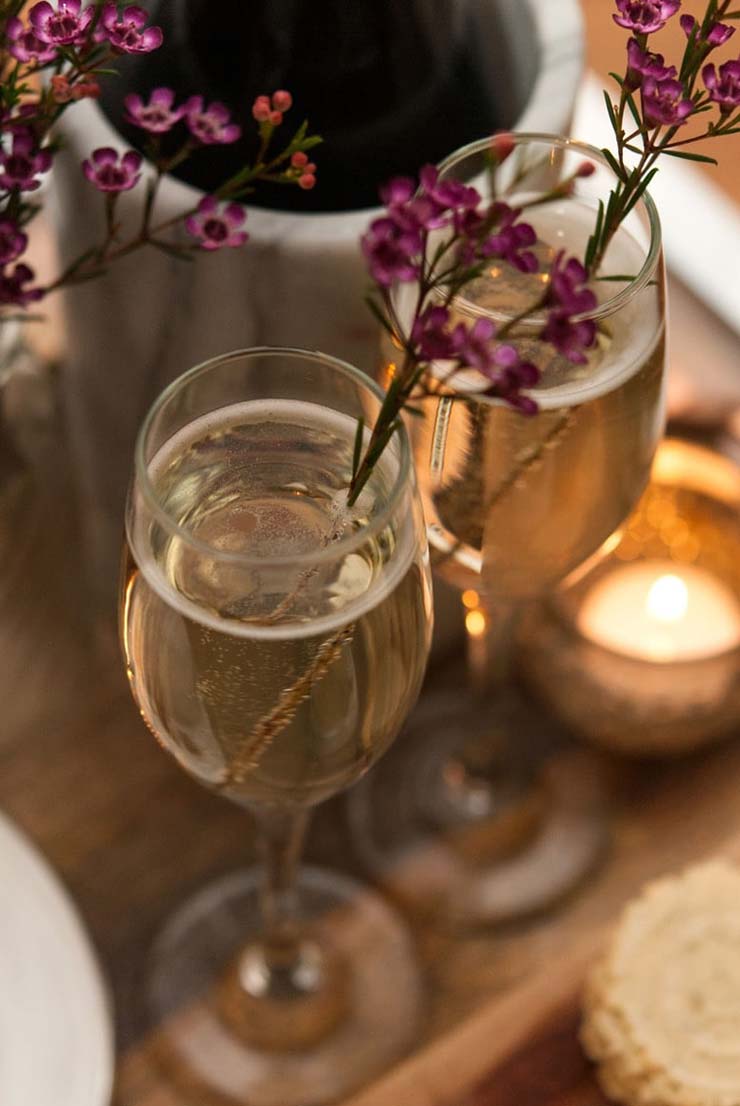 Take a look at these romantic valentine's day ideas for you and your love! Cocktails, flowers and a little cheese plate may be the perfect way to spend a romantic Valentine's evening at home. #cocktails, #cocktailparty, #flowers, #cheeseplate, #valentinesday #valentine 2 full champagne glasses, garnished with flowers next to a small candle on a wooden tray, next to a champagne bottle.