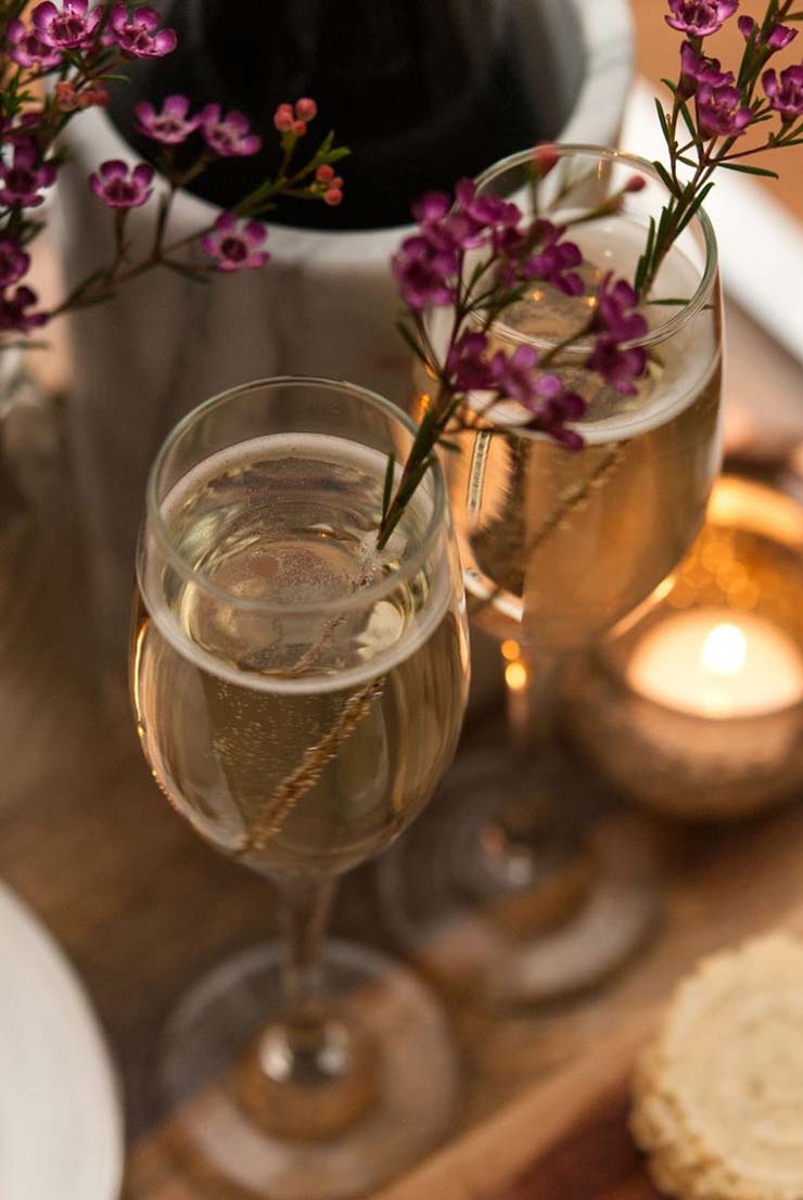 2 full champagne glasses, garnished with flowers next to a small candle on a wooden tray, next to a champagne bottle.