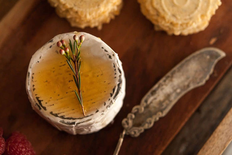 A cheese, drizzled in honey and garnished with a small flower beside an antique cheese knife and a few stacked crackers.