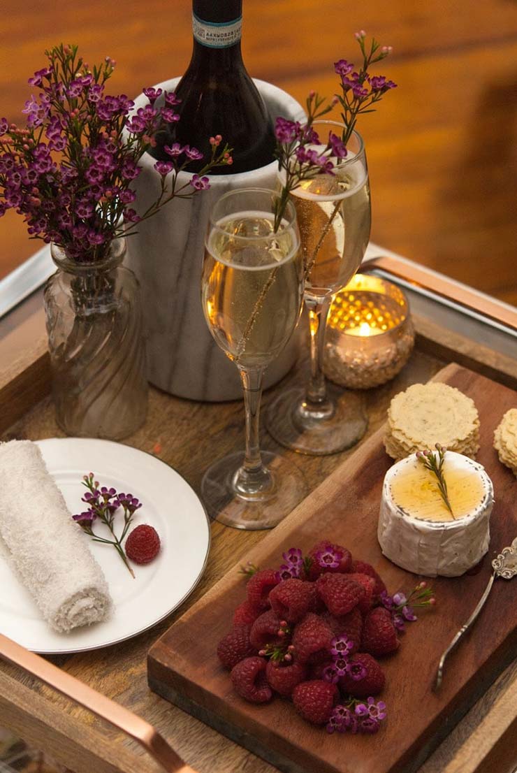 A wooden tray holding 2 champagne glasses with an assortment of cheeses, berries, crackers and flowers.