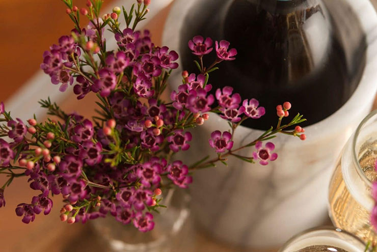 A glass vase of small flowers next to a marble wine holder with a bottle inside on a wooden tray.