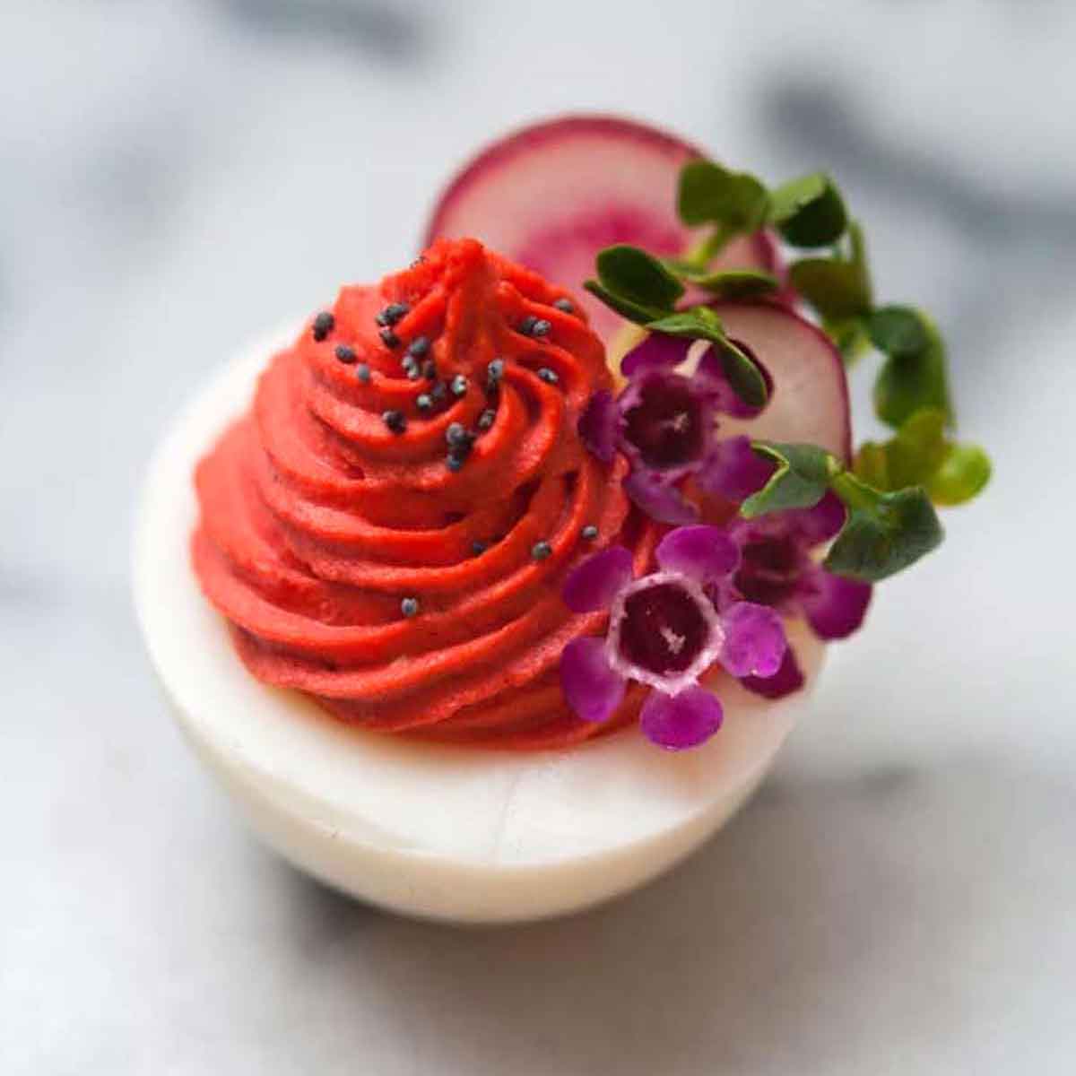 A red deviled egg, garnished with flowers and radishes on a marble slate.