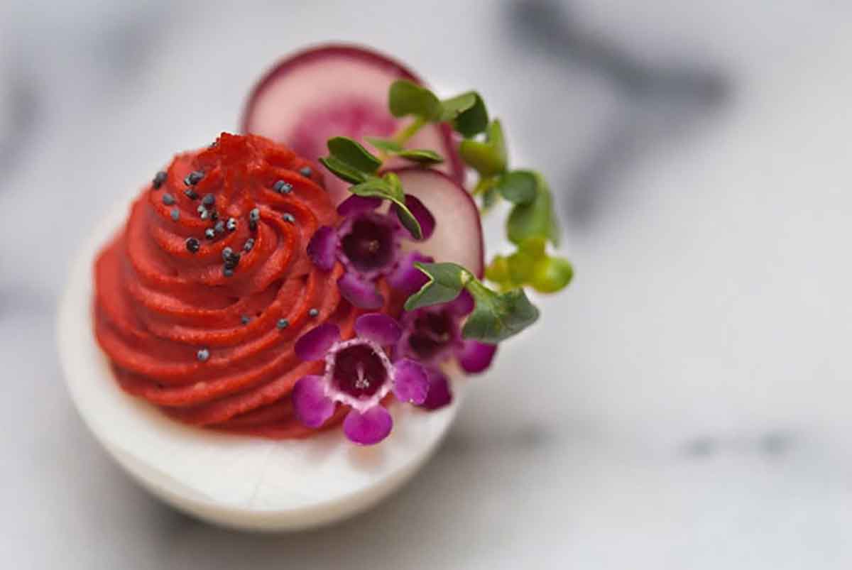 A deviled egg with red filling, garnished with sprouts, radishes, tiny flowers and poppyseeds on a white marble table.