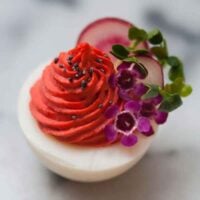 A deviled egg with red filling, garnished with sprouts, radishes, tiny flowers and poppyseeds on a white marble table.