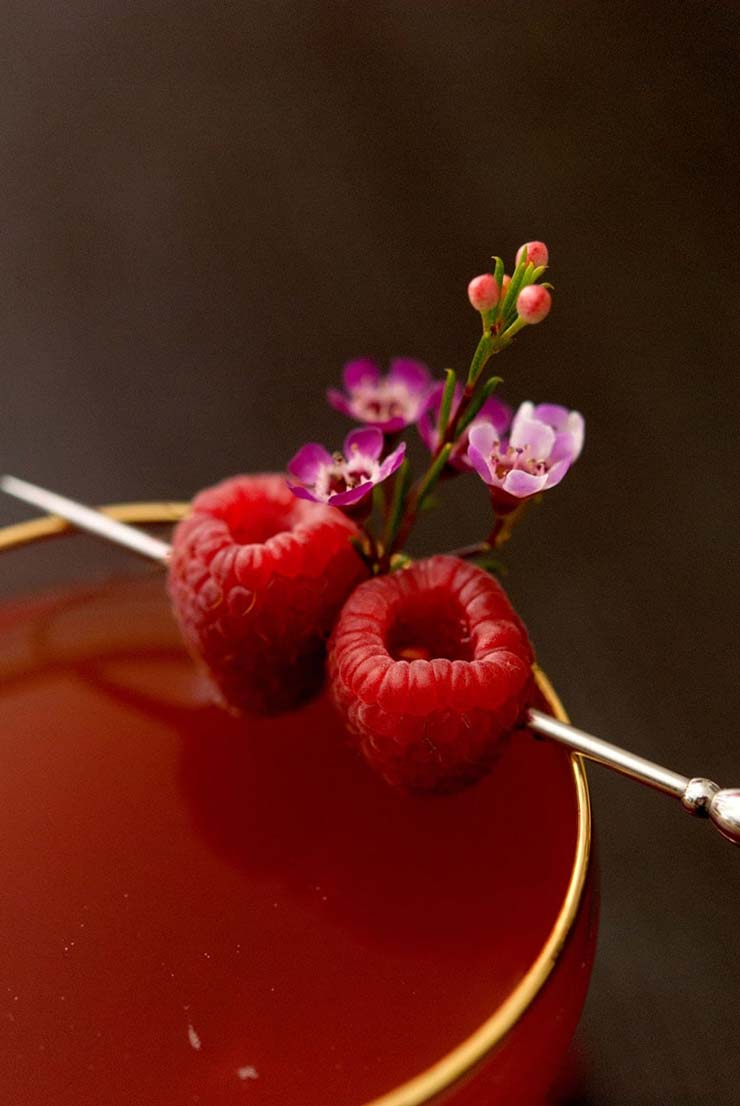 A closeup of a cocktail garnished with small lowers and two raspberries, pierced with a cocktail pin.