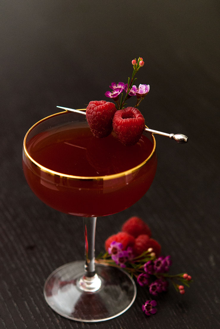 A cocktail on a black table garnished with raspberries and small flowers.