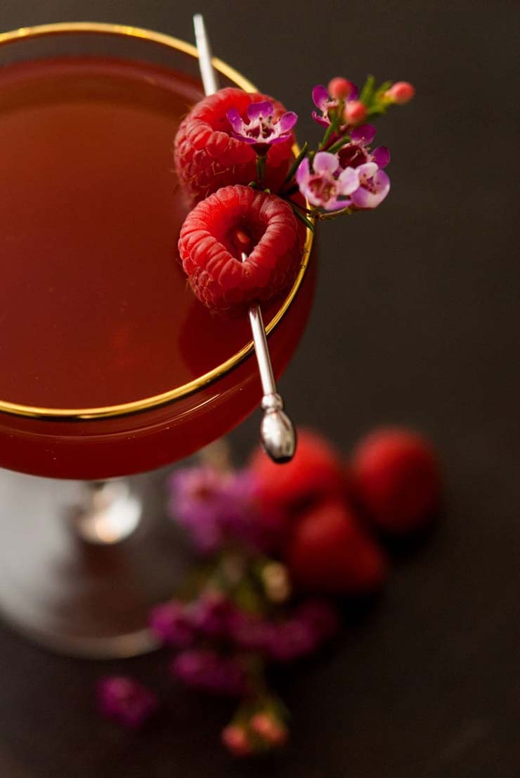 A cocktail garnish with small flowers and two raspberries, pierced with a cocktail pin on the side of a cocktail.
