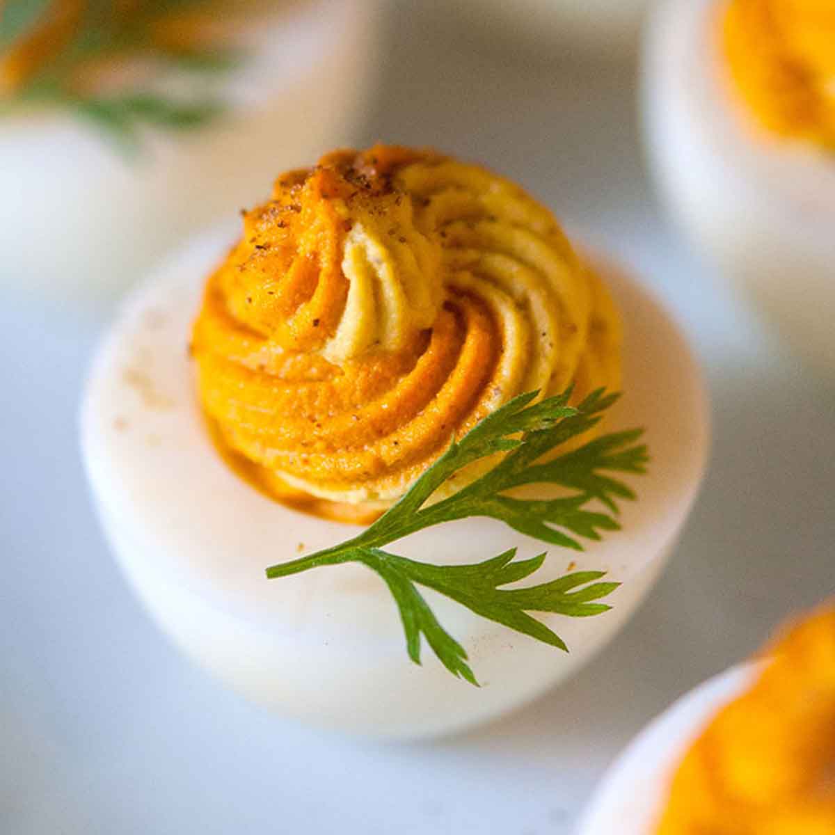 A closeup of a multi-colored deviled egg on a plate with a few others, garnished with a carrot frond.