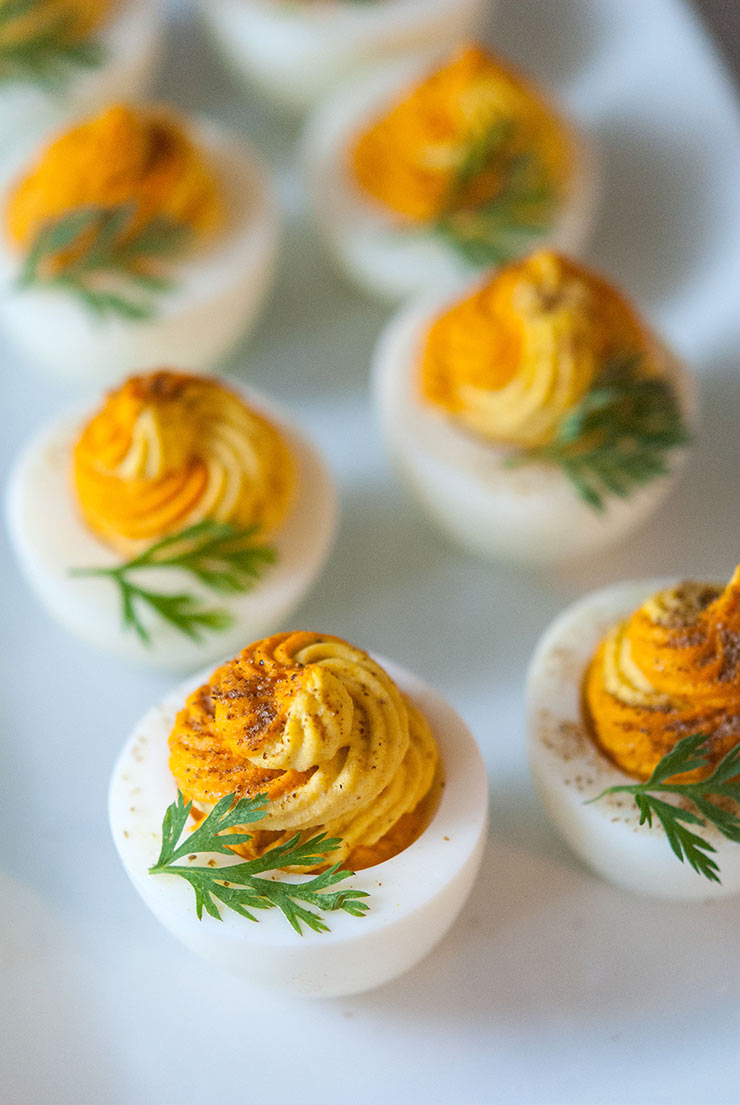 A closeup of a multi-colored deviled egg on a plate with a few others, garnished with a carrot frond.