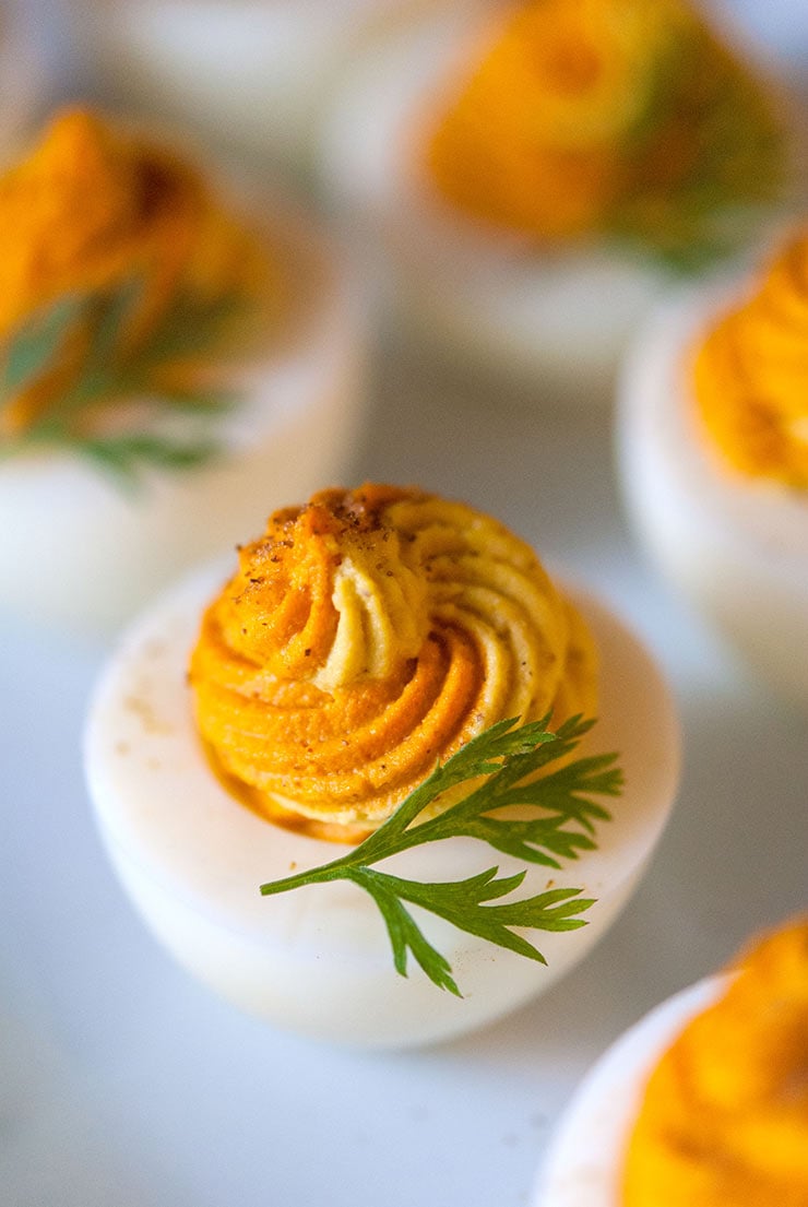 A closeup of a multi-colored deviled egg on a plate with a few others, garnished with a carrot frond.