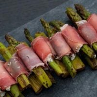 A pile of prosciutto-wrapped asparagus on a marble plate in a row.