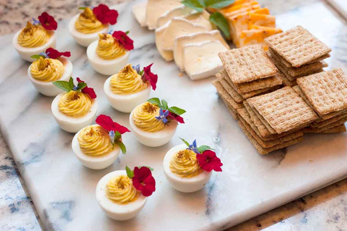 10 deviled eggs, garnished with flowers on a marble cheese board next to crackers and sliced cheese.